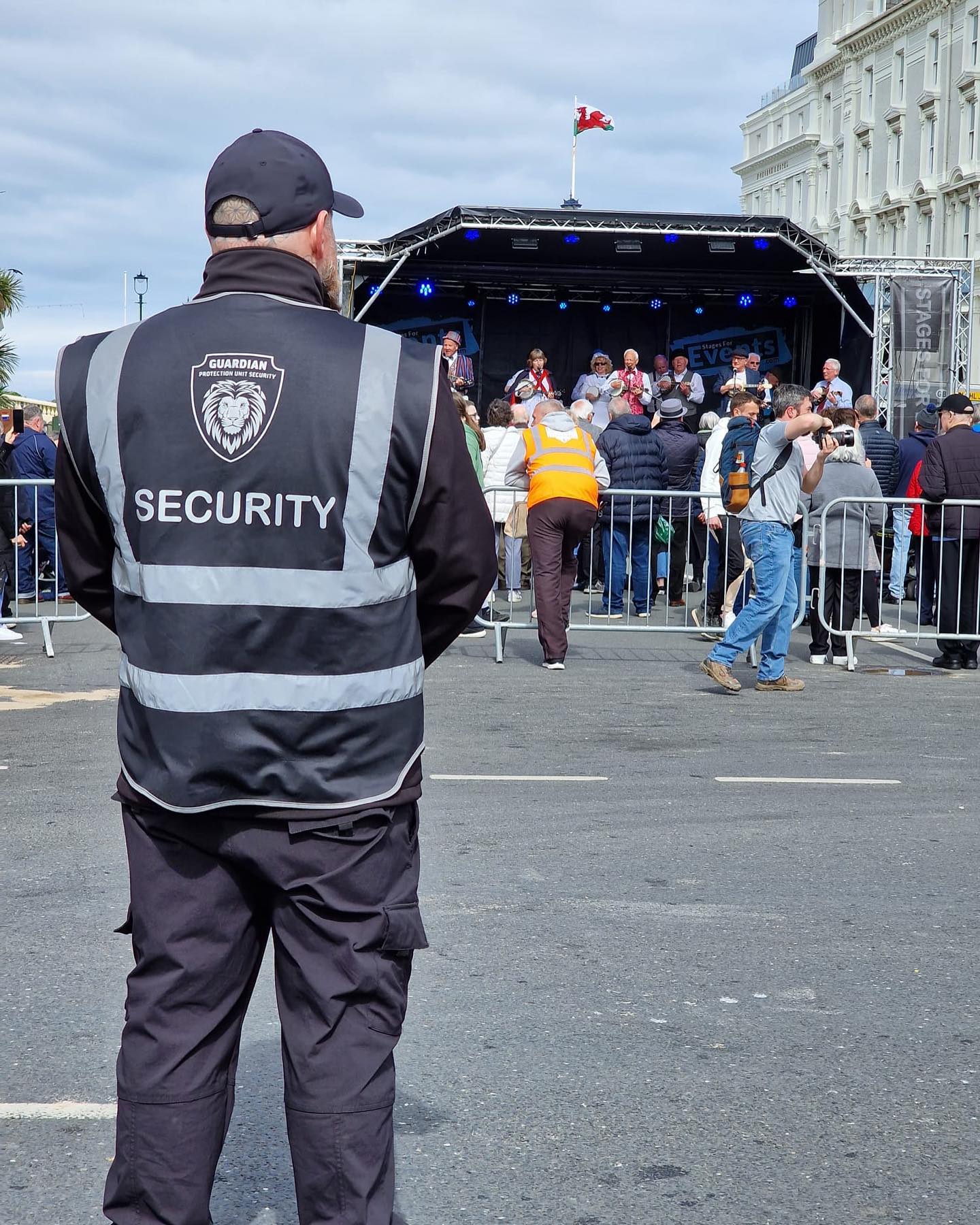 Security guard with reflective vest watching a crowd gathered in front of a stage where a group of elderly people is performing under a cloudy sky.