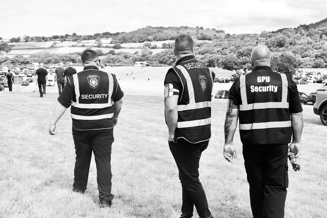 Three security personnel walking on a grassy field toward a countryside area with parked cars and hills in the background.