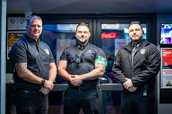 Three security guards standing side by side in uniform inside a building entrance.
