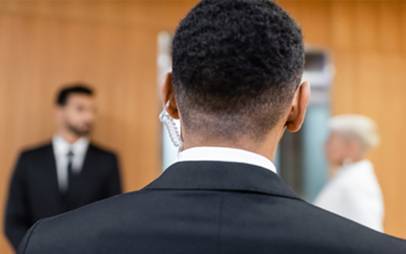 Close-up of a bodyguard wearing an earpiece standing with his back to the camera, facing a man and woman in business attire.