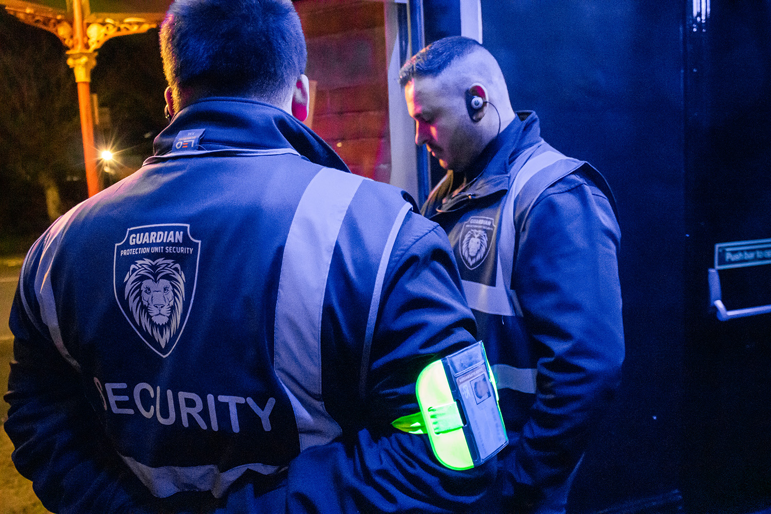 Two security guards in high-visibility jackets with a lion emblem and the word 'SECURITY' on the back, standing near a door at night.