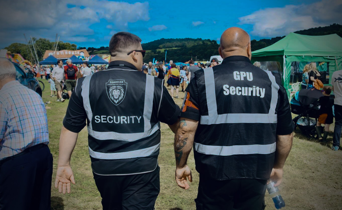 Two security guards wearing black vests walking at an outdoor event with tents and crowd in the background.