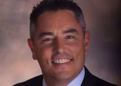 Smiling man with short dark hair wearing a dark suit and light blue shirt against a blurred background.
