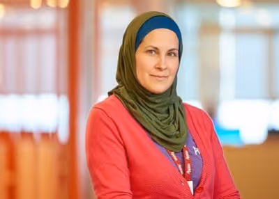 Woman wearing a green hijab and red cardigan, sitting and smiling in a softly lit indoor setting.