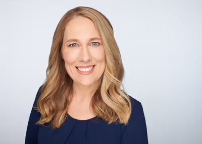 Smiling woman with shoulder-length blonde hair wearing a dark blue top against a light background.