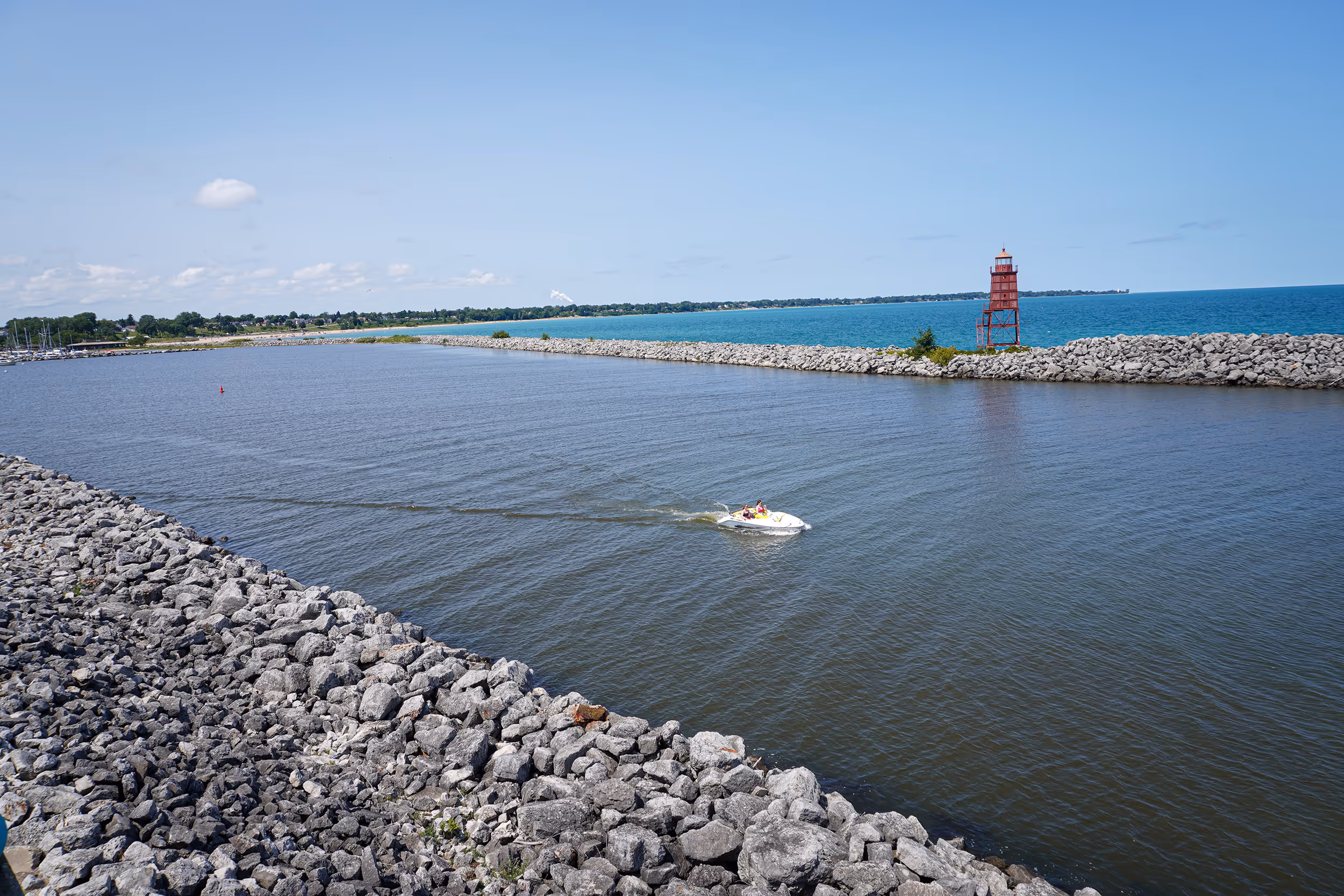 Small motorboat cruising on calm harbor water bordered by rocky breakwaters with a red lighthouse on the right side under a clear blue sky.
