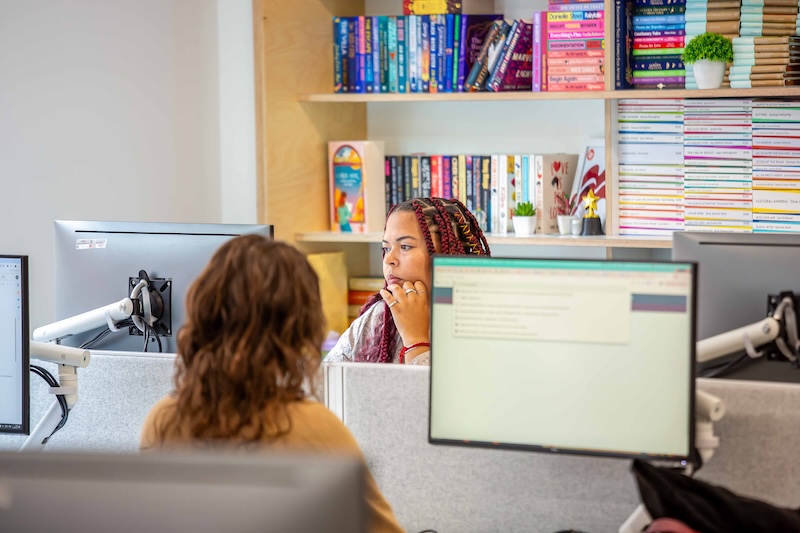 Two female Pan Macmillan employees working at computers in a warmly lit office. 