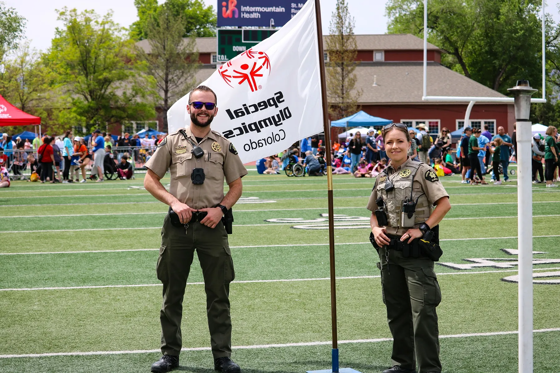 Two uniformed law enforcement officers standing on a sports field beside a flag that reads 'Special Olympics Colorado' with a crowd of people and trees in the background.