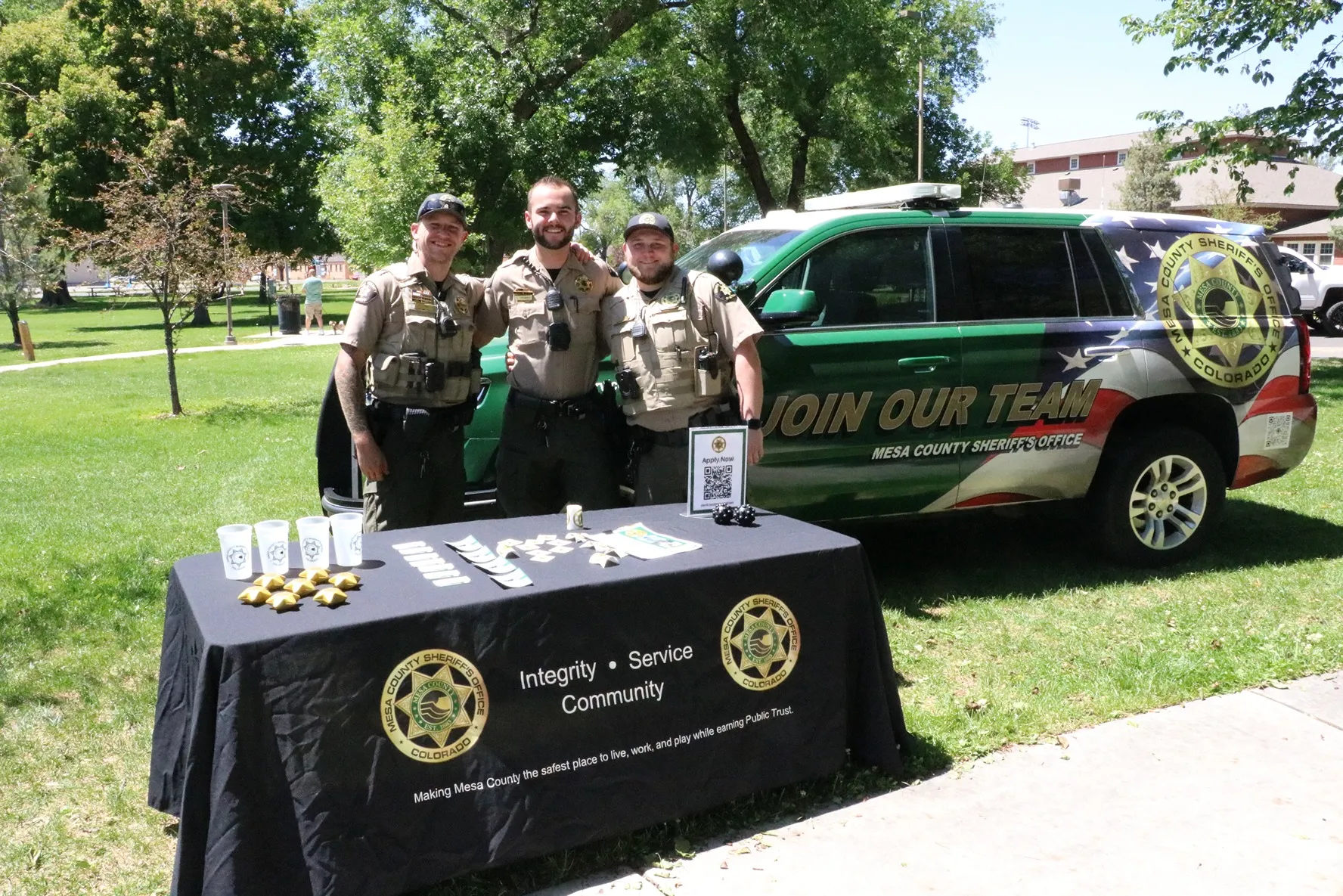 Three uniformed Mesa County Sheriff's deputies standing behind a recruitment table and green sheriff's vehicle with 'Join Our Team' signage in a park.