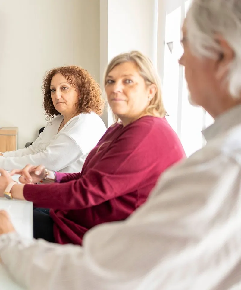 Tres personas mayores sentadas en círculo participando en una reunión o terapia grupal en un entorno interior iluminado.