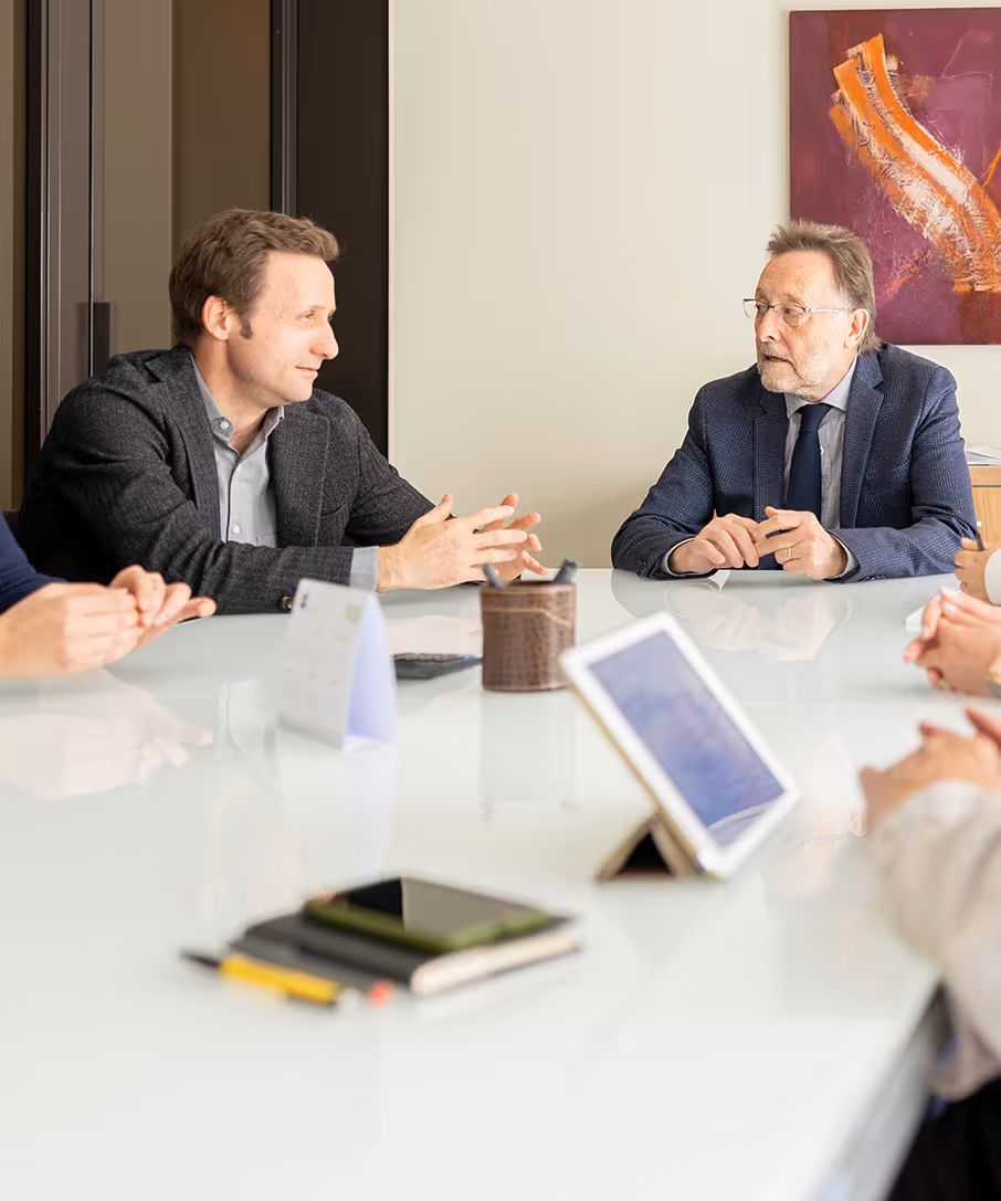 Dos hombres conversando en una reunión de oficina, sentados en una mesa blanca con una tablet y cuadernos.