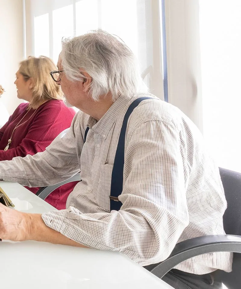 Hombre mayor con gafas y tirantes blancos sentado en una mesa blanca junto a una mujer con suéter burdeos en una sala iluminada.