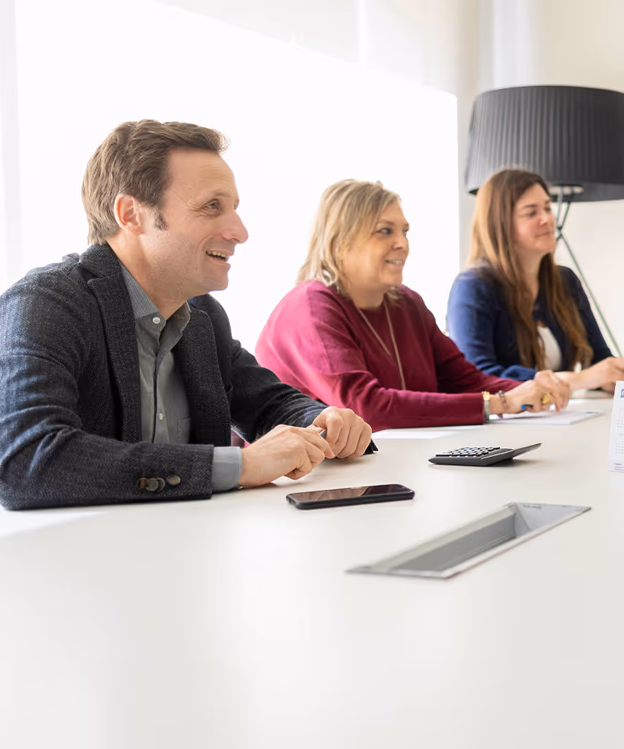 Tres personas sentadas en una mesa de oficina, mirando hacia adelante y sonriendo durante una reunión.