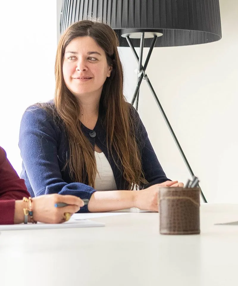Mujer sonriente de cabello largo sentado en mesa blanca con lámpara de pie negra al fondo.