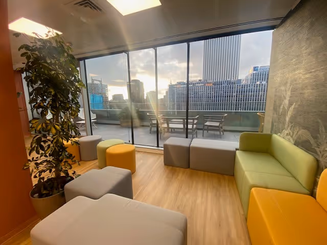 Oficina moderna con asientos modulares de colores, suelo de madera y grandes ventanales con vista a la ciudad al atardecer.