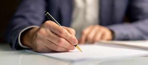 Mano escribiendo con bolígrafo negro con detalles dorados sobre papel blanco.
