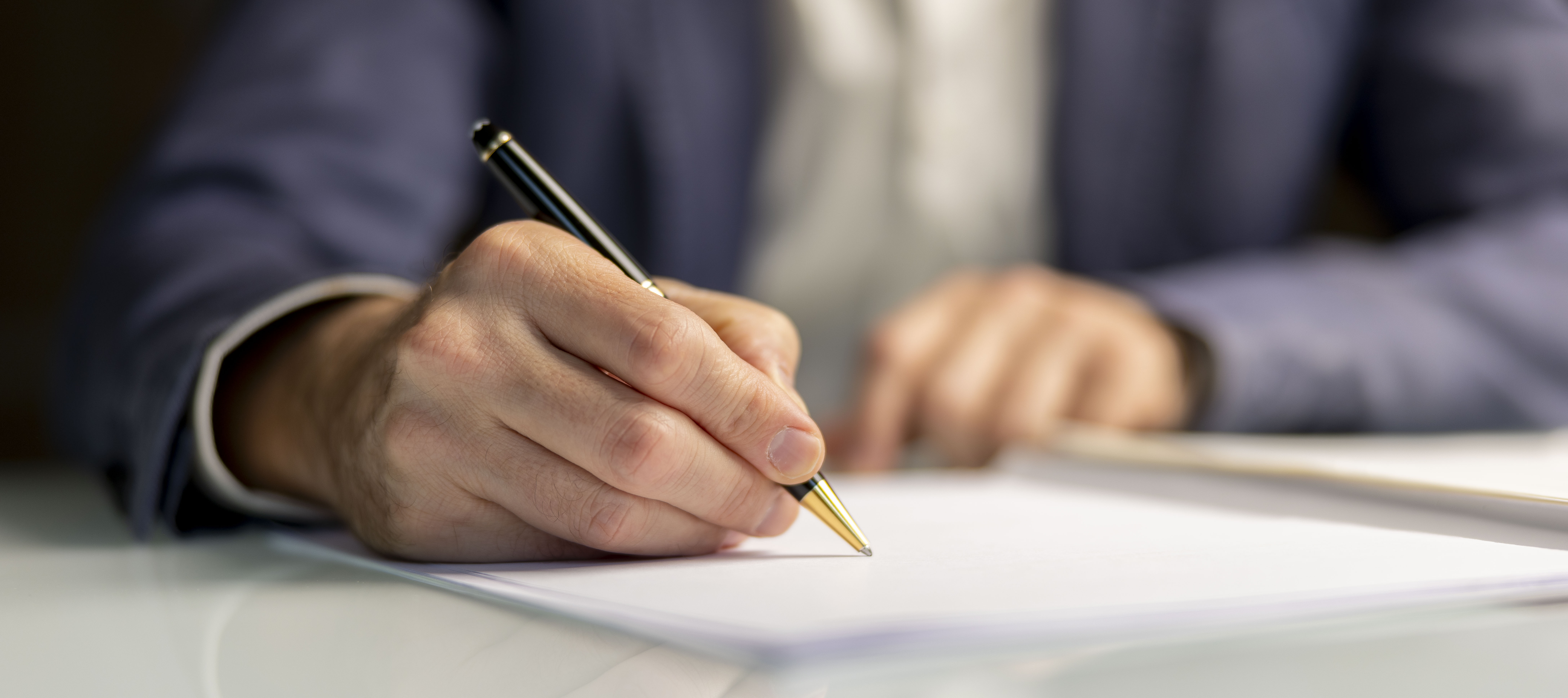 Mano escribiendo con bolígrafo negro con detalles dorados sobre papel blanco.