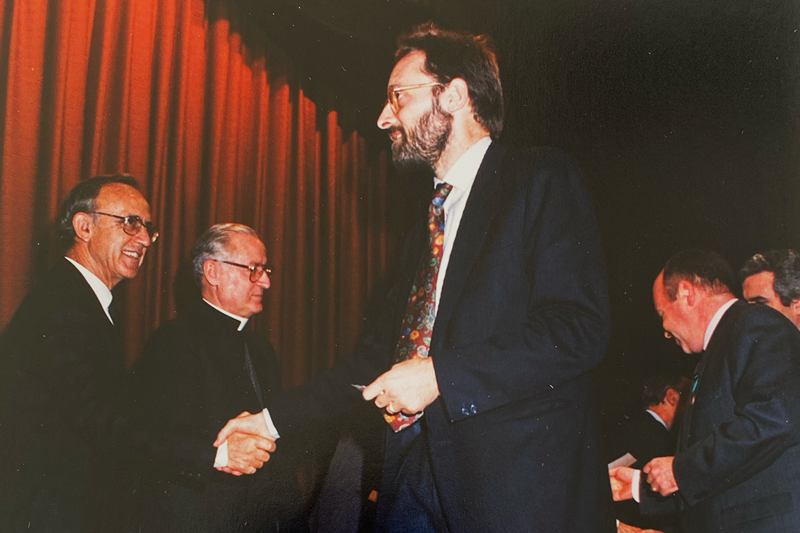 Hombres de traje saludándose con un apretón de manos frente a una cortina roja, uno con sotana de sacerdote.