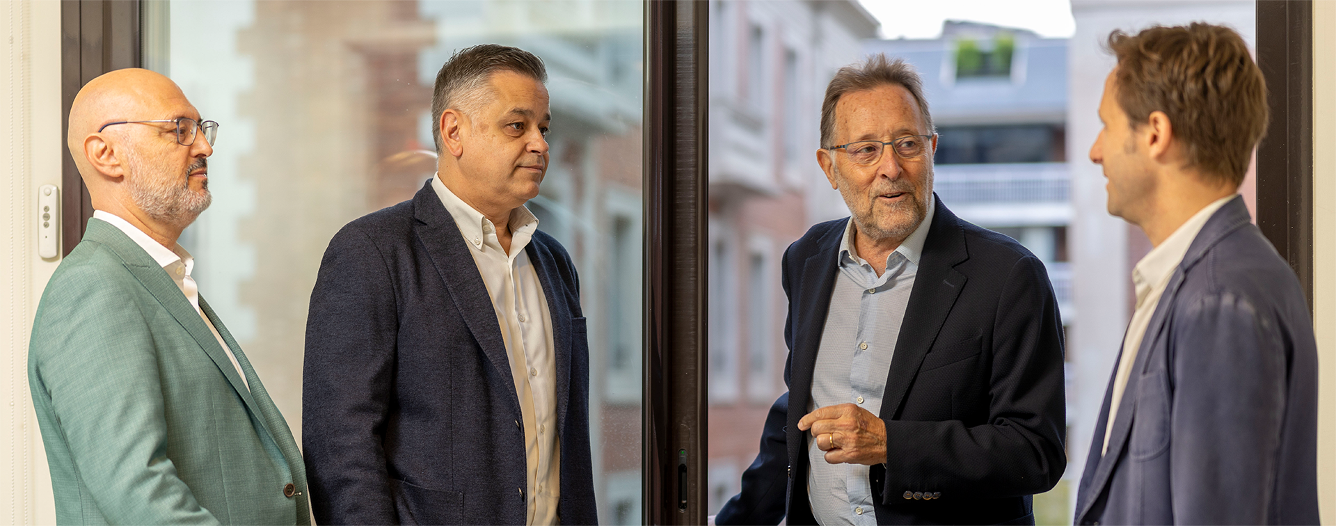 Cuatro hombres en traje conversando junto a una ventana grande con vista a edificios urbanos.