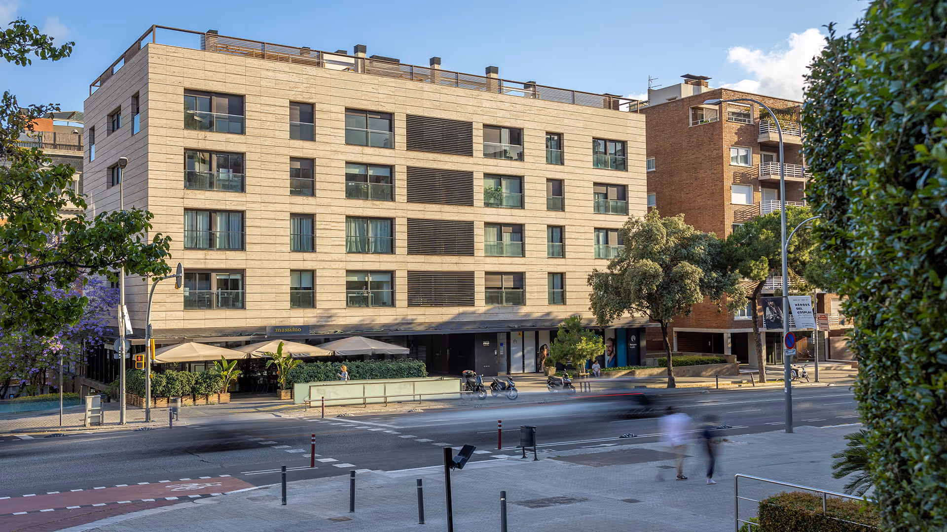 Edificio moderno de cuatro pisos con terraza y terraza exterior con sombrillas en una avenida urbana con árboles y personas caminando.