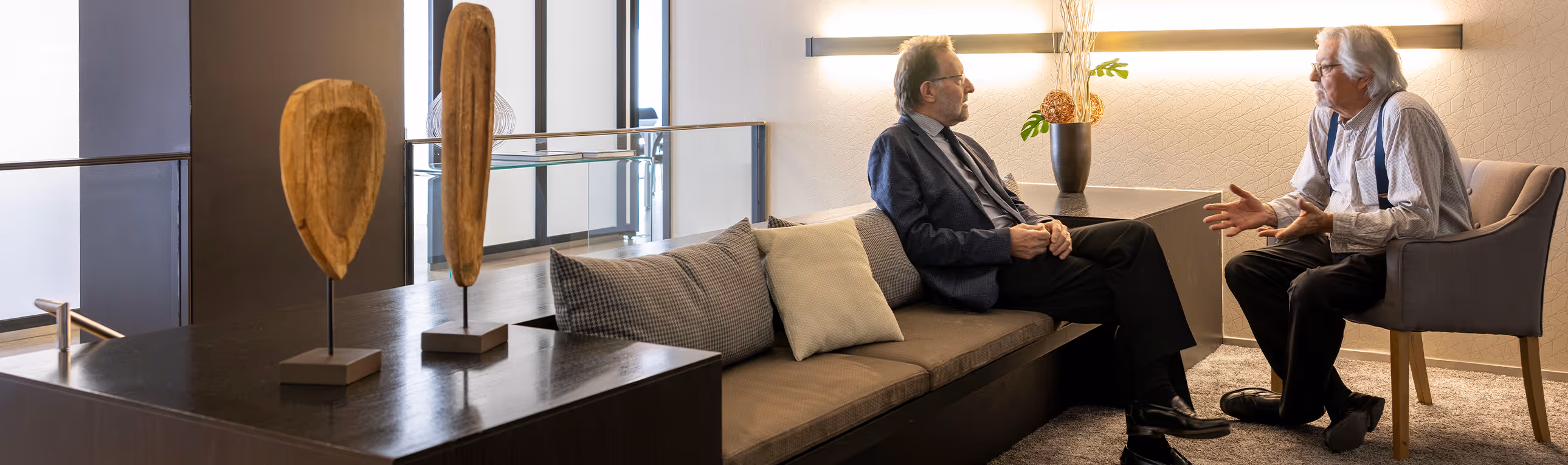 Dos hombres mayores conversando sentados en una sala moderna con sofá, silla y decoración de madera.
