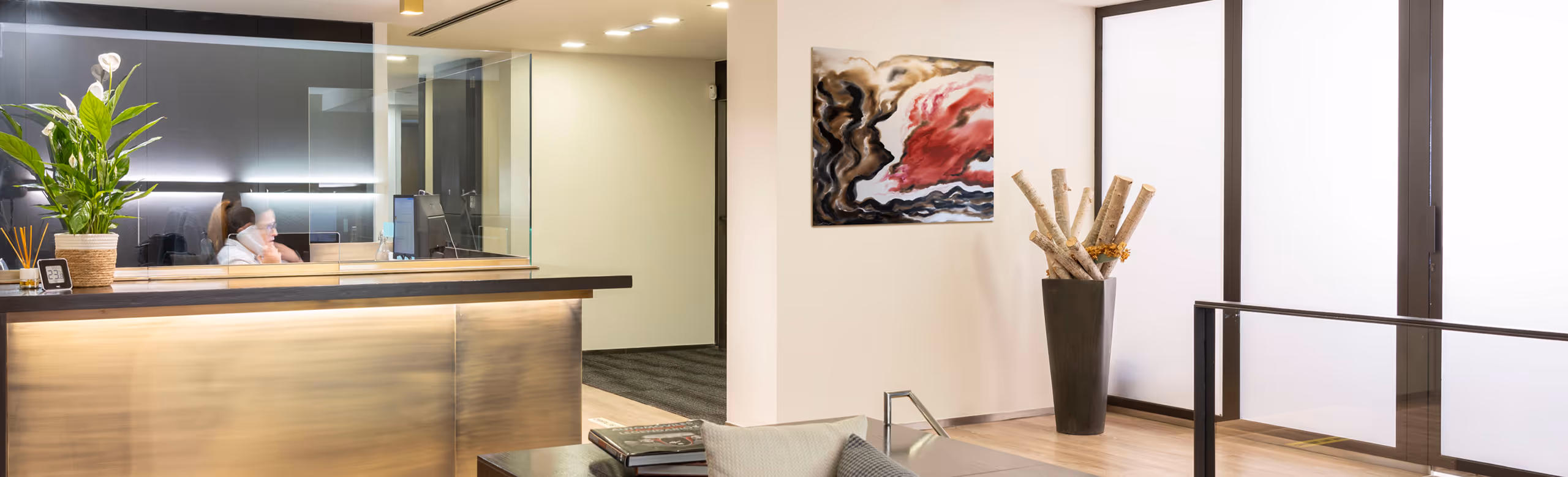 Recepción moderna con mostrador iluminado, planta verde, persona hablando por teléfono y decoración abstracta en la pared.