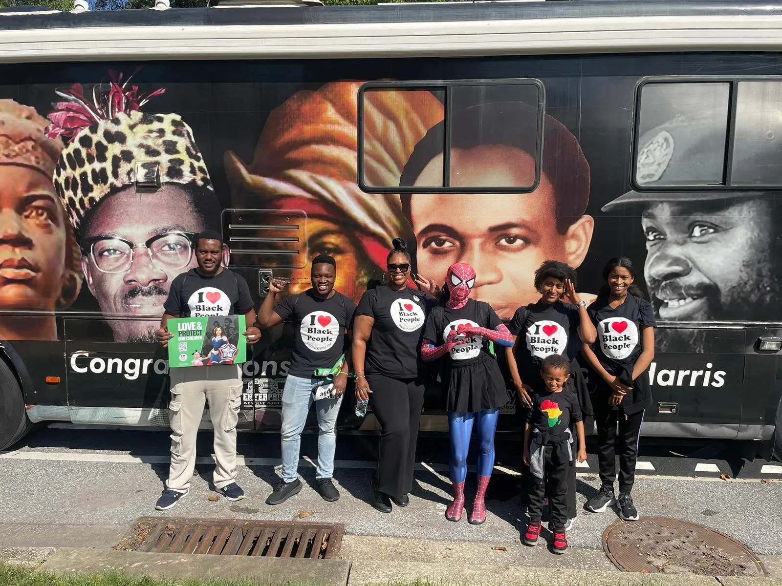 Group of seven Black people, including a child and one dressed as Spiderman, standing and smiling in front of a bus with large portraits of influential Black figures.