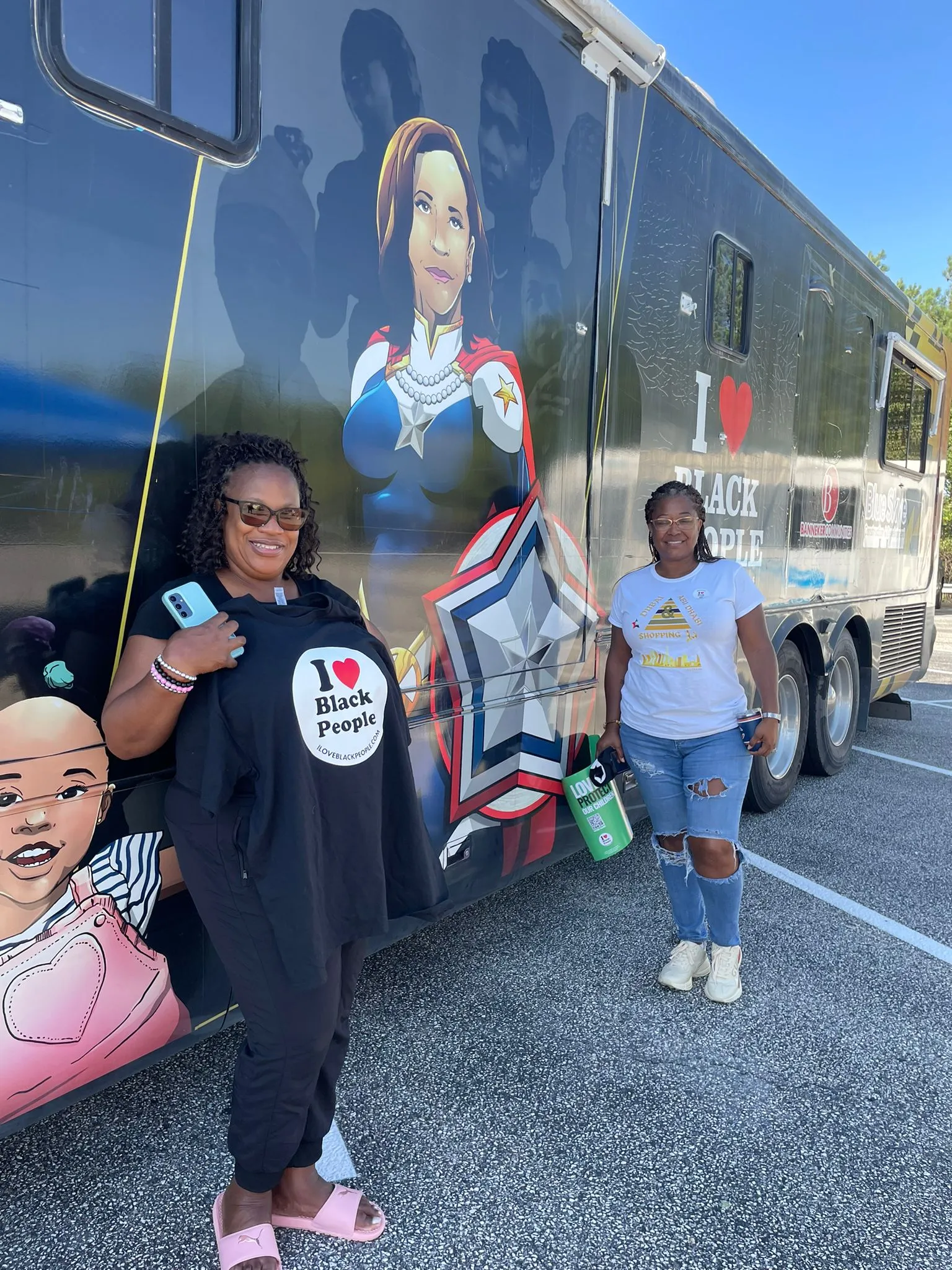 Two women standing in front of a vehicle with illustrations and text saying 'I ❤️ Black People,' one holding a matching black shirt and the other holding a green water bottle.