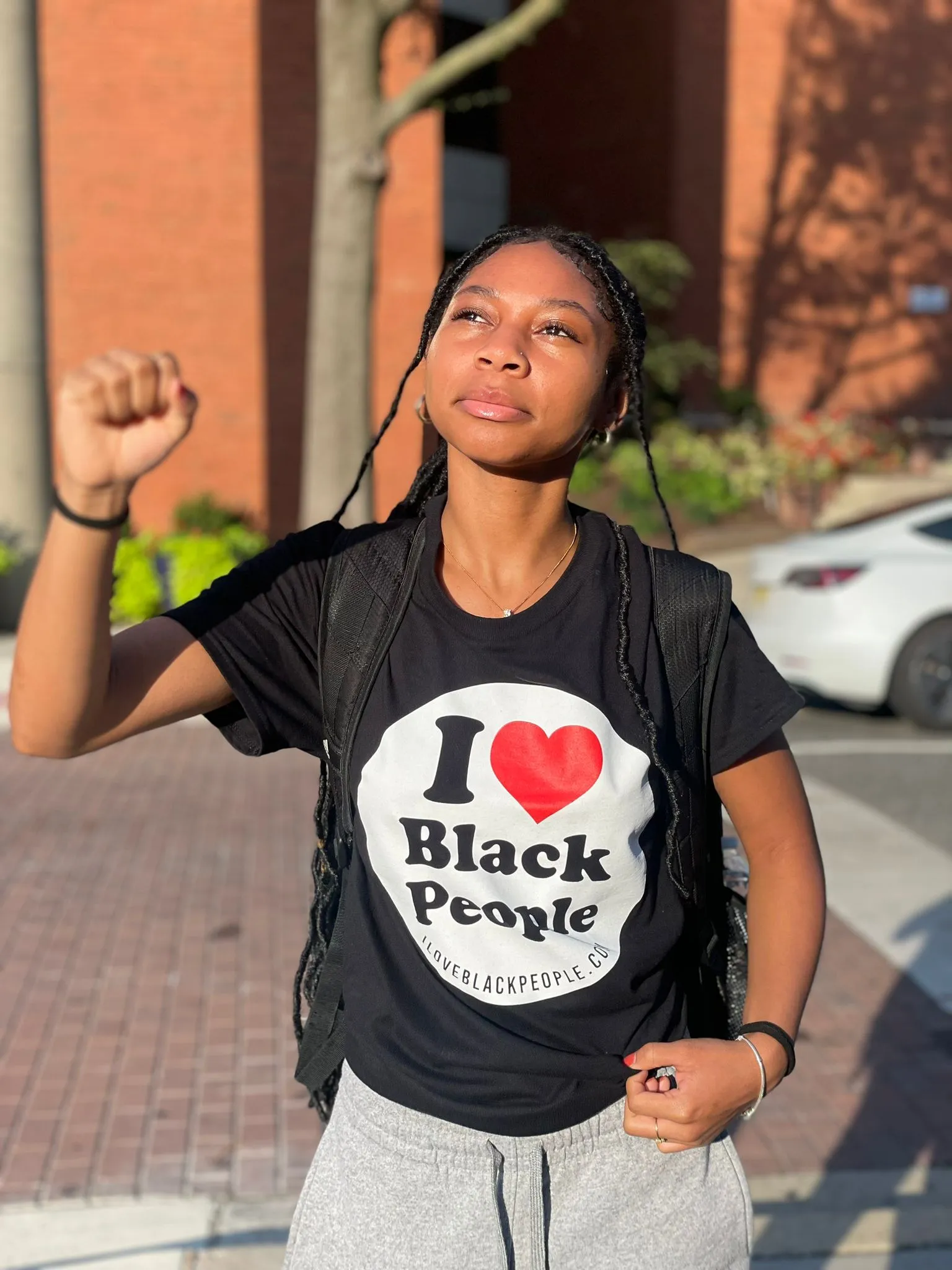 Young woman with braided hair wearing a black T-shirt that reads 'I ♥ Black People' raising her fist outdoors in sunlight.