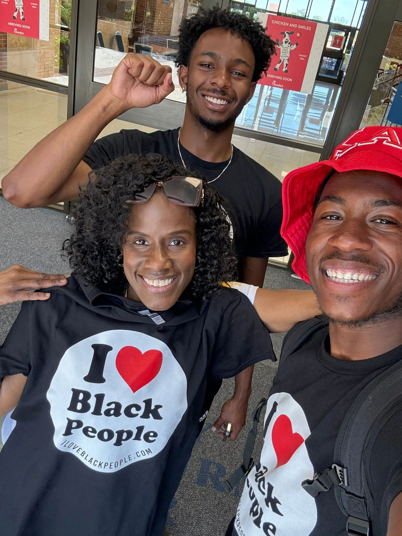 Three smiling Black individuals, two wearing matching black "I ♥ Black People" shirts, posing indoors with one person raising a fist.