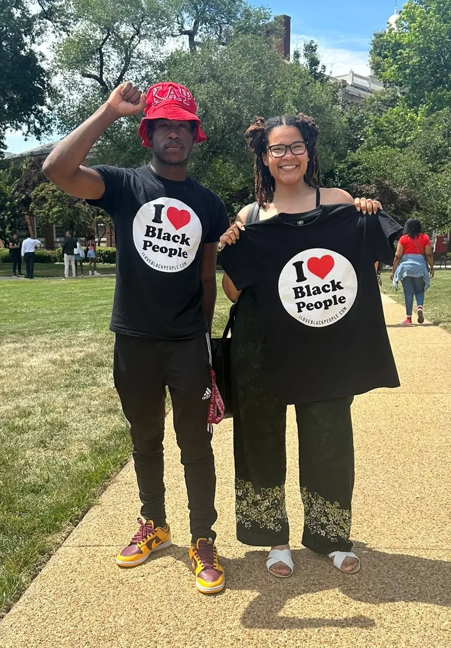 Two people outdoors, one wearing a red hat and black t-shirt with 'I love Black People' text holding a fist up, the other smiling and holding a matching t-shirt.