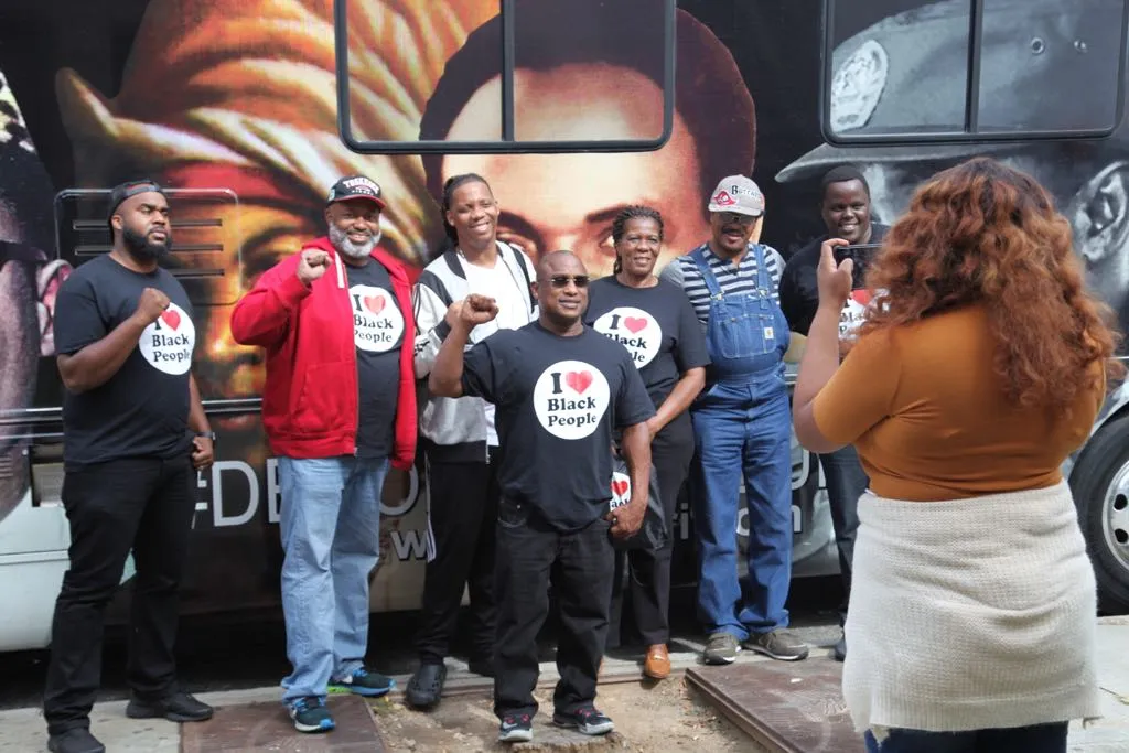 Group of six Black men and one Black woman posing with raised fists, wearing 'I love Black People' shirts, while a woman takes their photo in front of a mural.