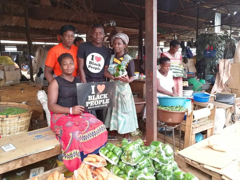 Group of people at a market with fresh vegetables; one person holds a sign reading 'I love Black People #decoloniseyourlife'.