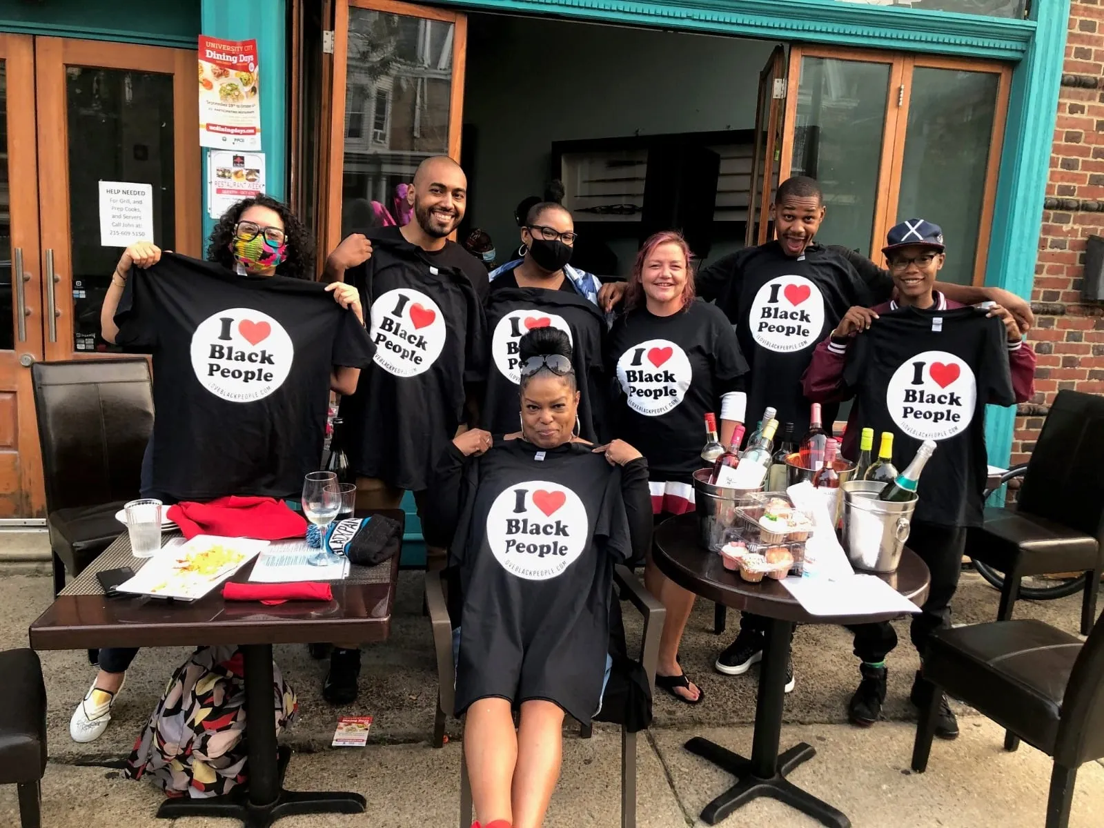Group of eight people smiling and holding black t-shirts that say 'I ❤️ Black People' outside a restaurant.