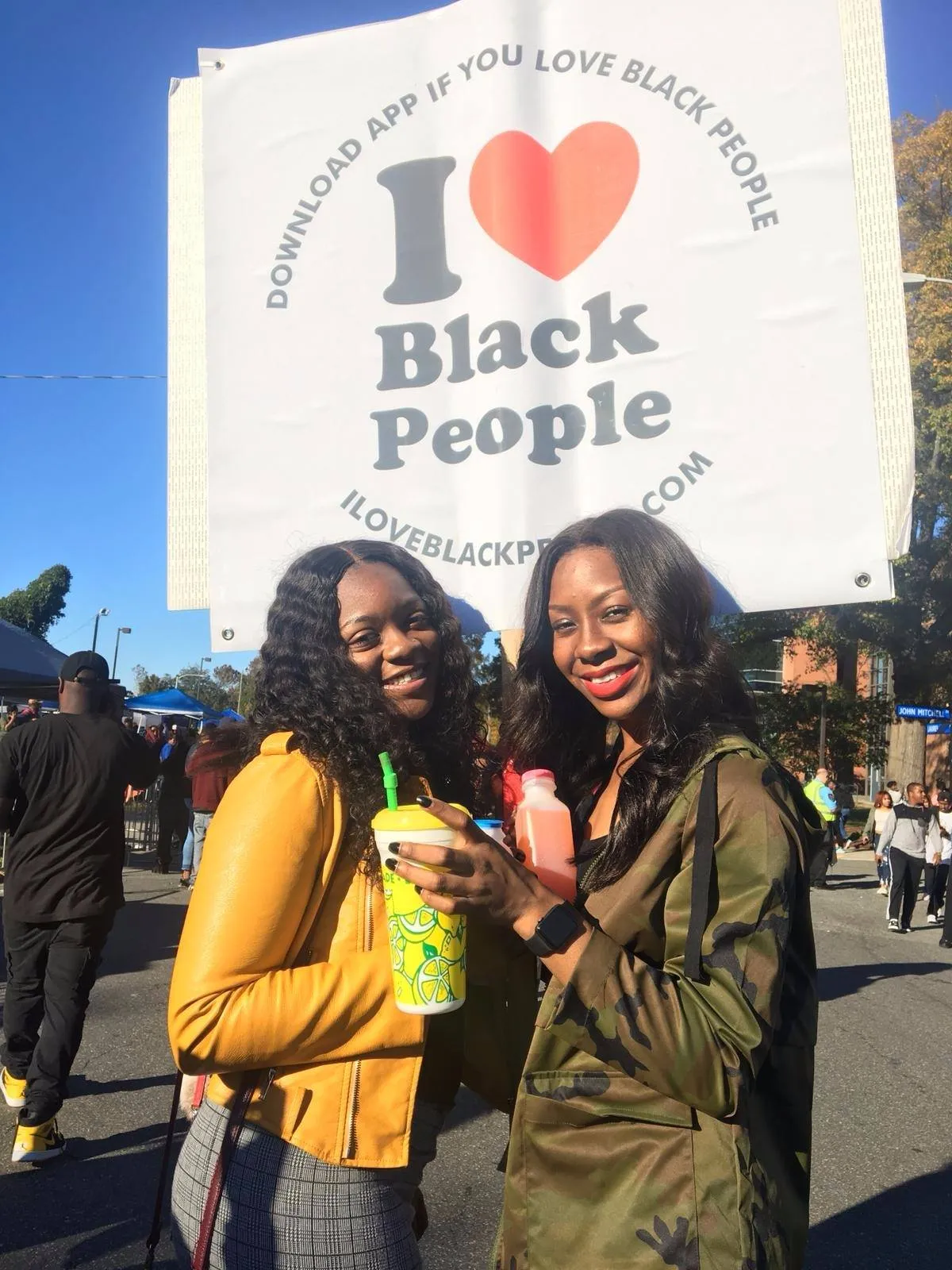 Two smiling Black women holding drinks stand in front of a sign reading 'I love Black People' at an outdoor event.