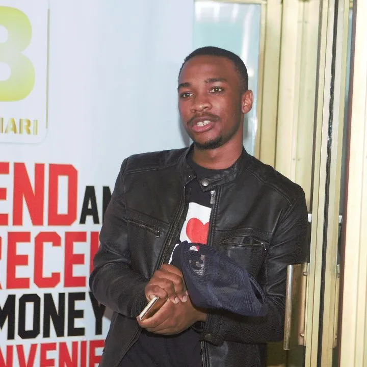 Man wearing a black leather jacket and holding a smartphone and a cap, standing indoors near a glass door with partially visible red and black text on the wall behind him.