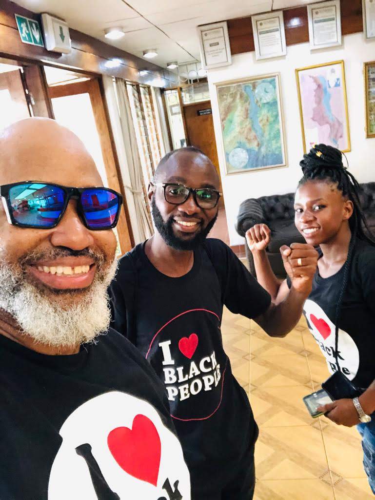 Three smiling Black people wearing black shirts with 'I love Black people' text, posing with raised fists indoors.