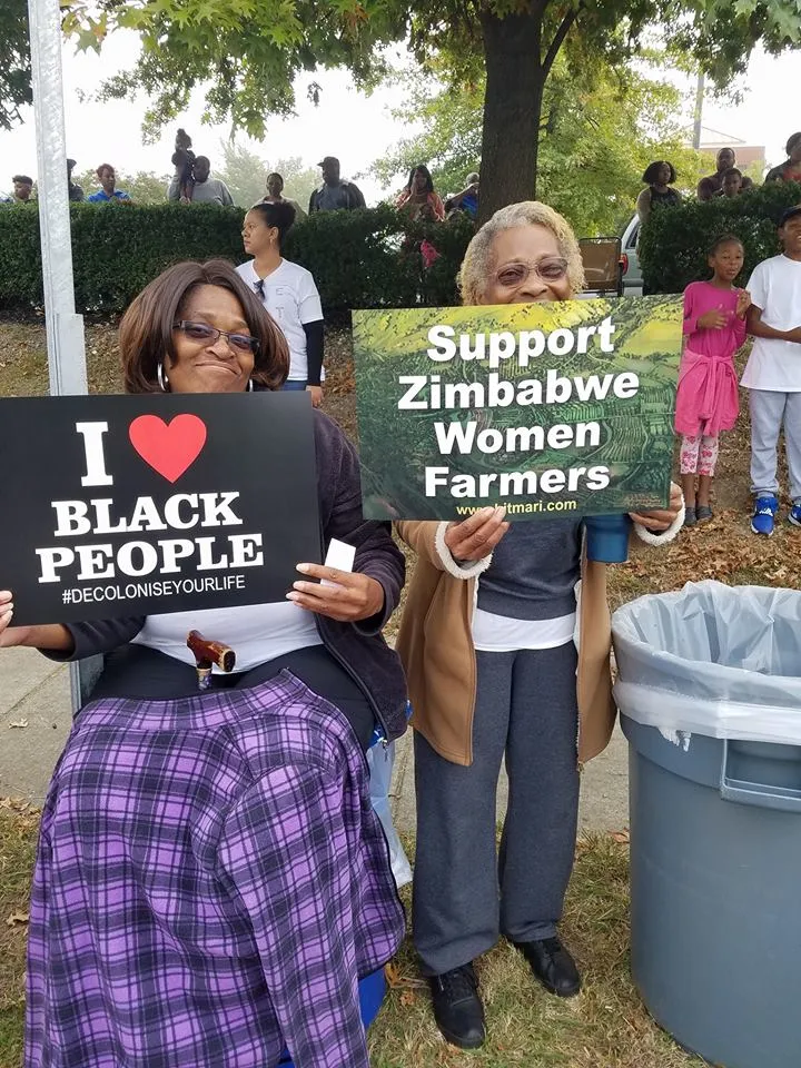 Two women holding signs: one says 'I love Black People #DecoloniseYourLife' and the other says 'Support Zimbabwe Women Farmers' at an outdoor event.