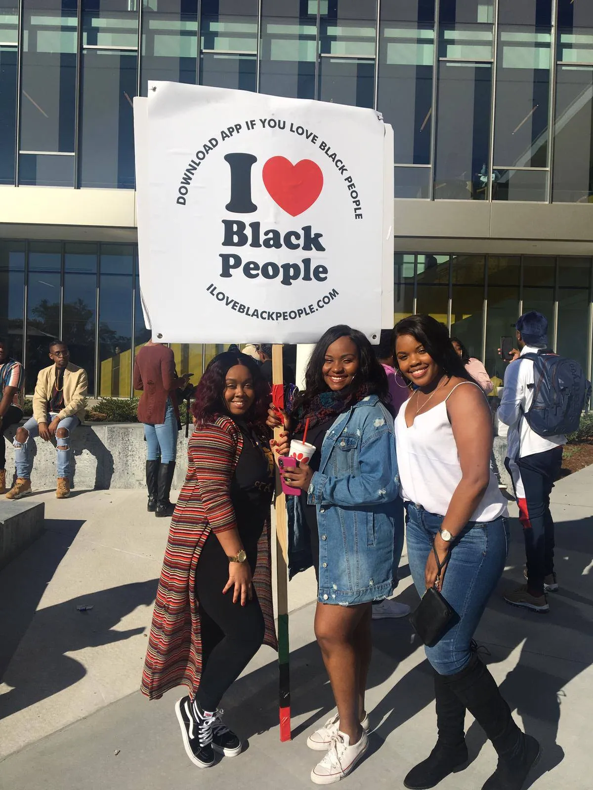 Three smiling Black women standing outdoors in front of a building, holding a sign that says 'I love Black People' with a red heart.
