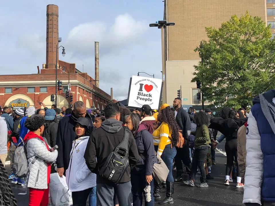 Crowd of people gathered outdoors near buildings with one person holding a sign that says 'I love Black People'.