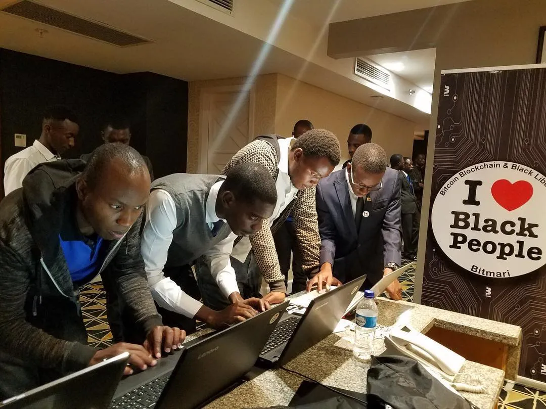 Group of men focused on typing at laptops in an indoor event with a sign reading 'I love Black People' in the background.
