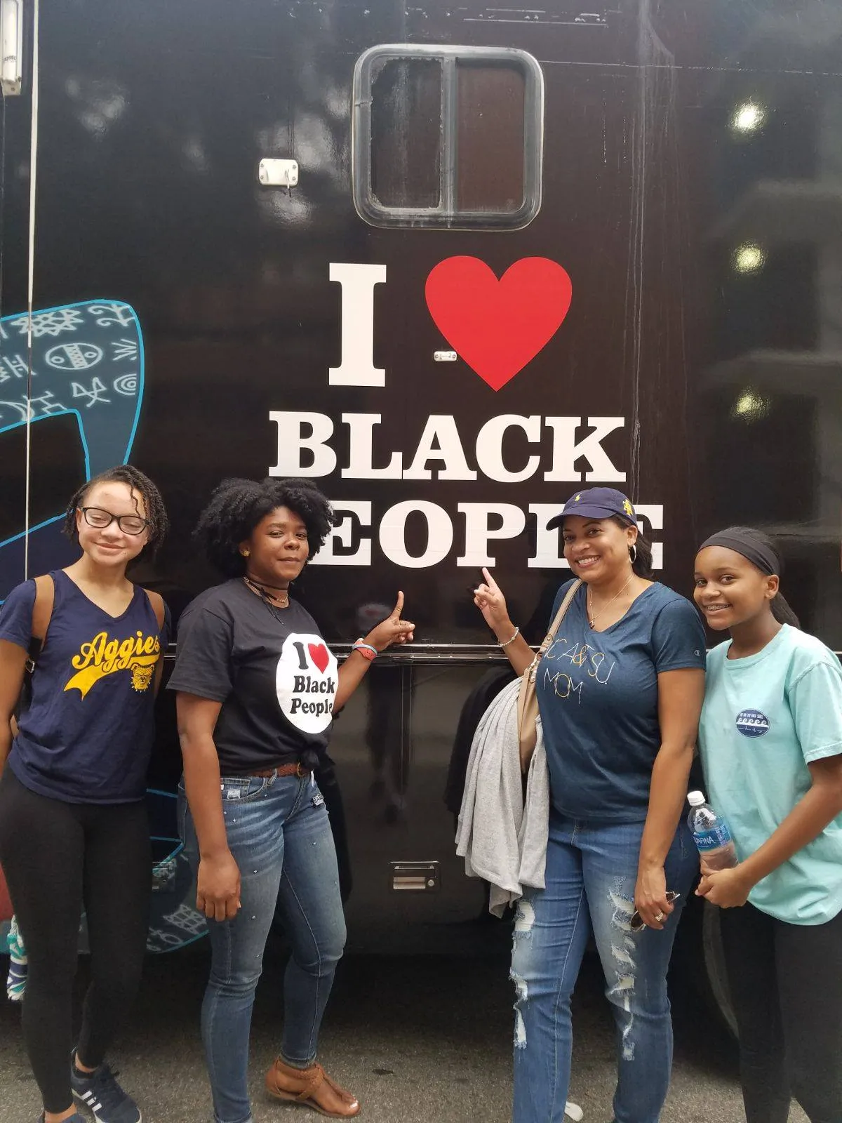 Four smiling Black women standing in front of a black vehicle with the text 'I ♥ Black People', two pointing at the text.