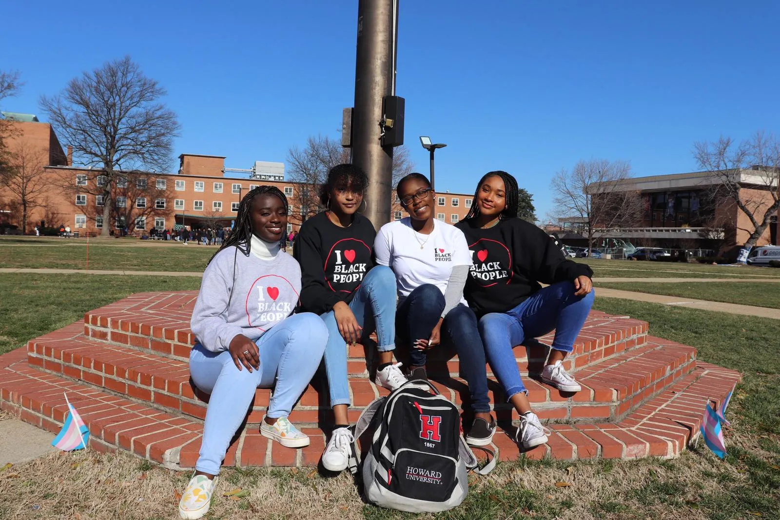 Four young women wearing 'I heart Black People' shirts sitting on brick steps outdoors with a Howard University backpack in front.