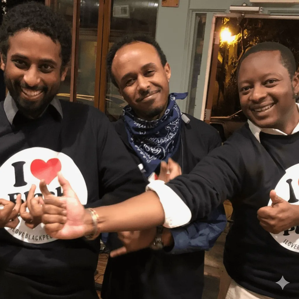 Three men smiling indoors, two wearing black sweatshirts with a red heart and the text 'I love black people'.