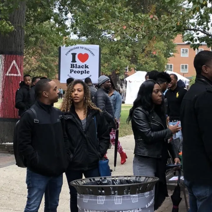 A group of people outdoors with a sign in the background reading 'I heart Black People' held up among trees and buildings.