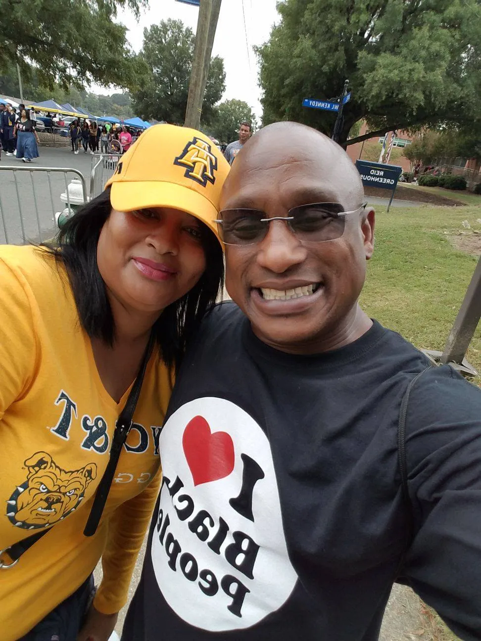 Smiling man and woman taking a selfie outdoors at an event, with the man wearing glasses and a black shirt that says 'I love Black People' and the woman wearing a yellow hat and shirt with a bulldog logo.