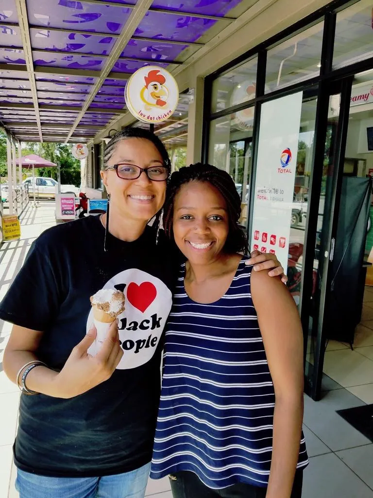 Two smiling women standing close together outside a storefront, one holding an ice cream cone and wearing a black 'I love black people' t-shirt.