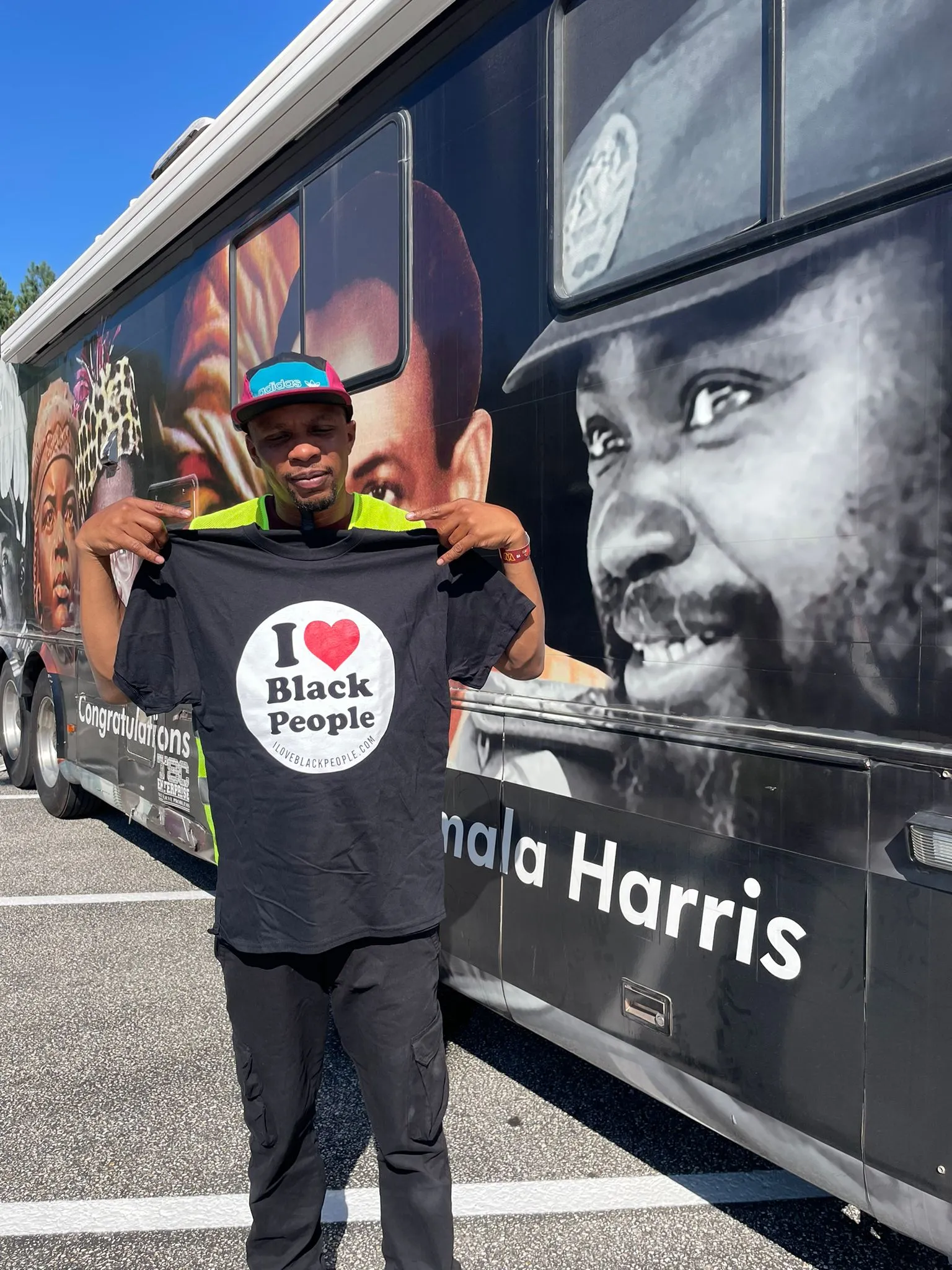 A man wearing a colorful cap holds up a black T-shirt that says 'I ❤️ Black People' in front of a bus with large portraits and text including 'Kamala Harris'.
