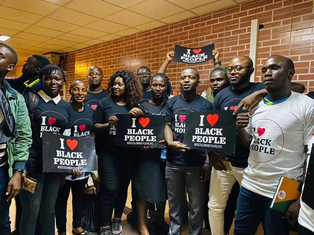 Group of Black people standing indoors holding signs and wearing shirts that say 'I love Black people' with a red heart and the hashtag #decoloniseyourlife.