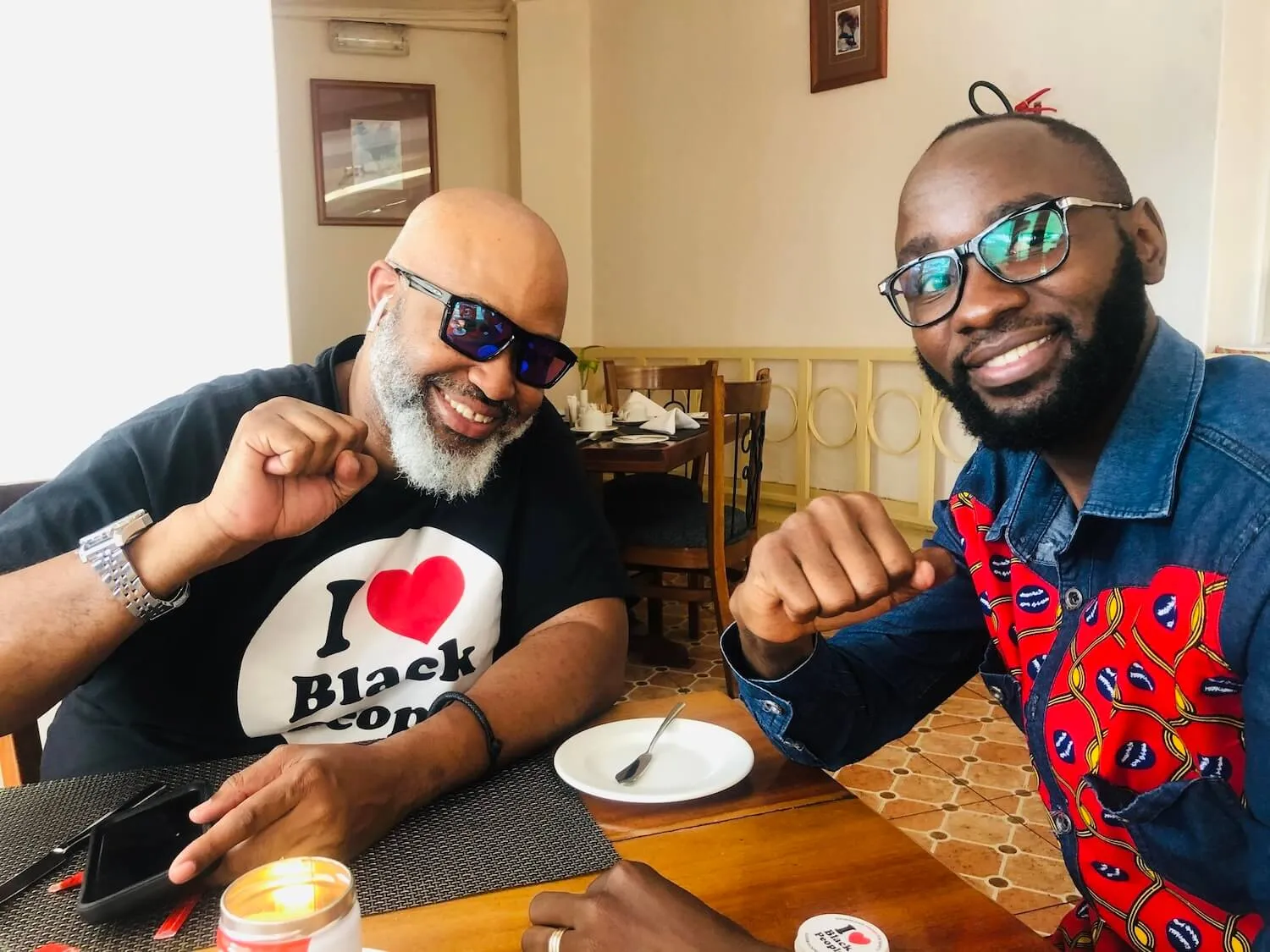 Two smiling men sitting at a restaurant table with fists raised in a friendly gesture; one wearing sunglasses and a black shirt with 'I ❤️ Black Co' text, the other wearing glasses and a colorful patterned shirt.